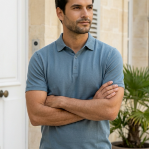 Homme les bras croisés portant un polo bleu ardoise coupe classique avec un pantalon beige, posant en extérieur.