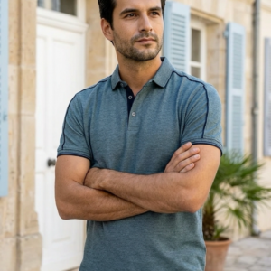 Homme les bras croisés portant un polo bleu gris chiné avec liserés bleu marine, coupe classique, et un pantalon bleu marine.