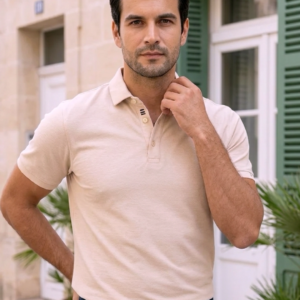 Homme portant un polo beige rosé coupe classique et un pantalon bleu marine, devant une façade claire.